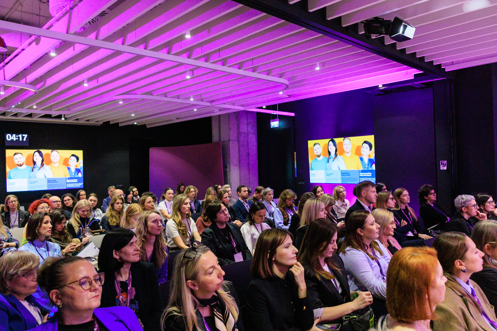 Publiczność siedzi w sali podczas gali Karty Różnorodności. Na ekranach widać zdjęcia uczestników wydarzenia. Oświetlenie sali w odcieniach fioletu.